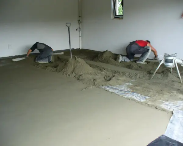 Dwóch budowlańców pracuje przy wylewce podłogi. W tle widać maszynę, która wygląda jak **mixokret**.