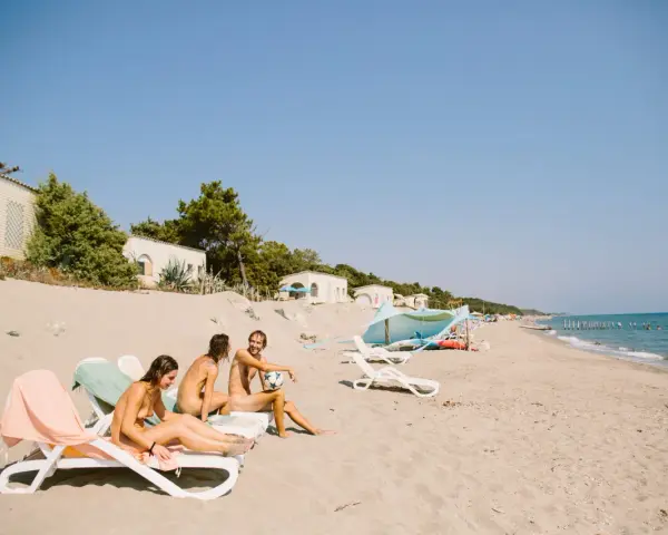 Trzy osoby relaksują się na plaży w nudystycznym obozie, siedząc na leżakach. Jeden z nich trzyma piłkę.
