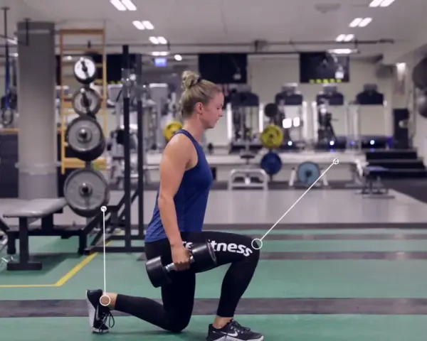 Kvinna utför utfallssteg med vikt i ett gym. Hon håller en hantel i ena handen och har ett ben framåt och ett bakåt.