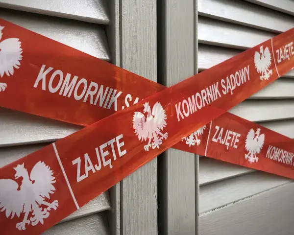 Czerwona taśma z napisem "KOMORNIK SĄDOWY" i godłem Polski krzyżuje się na szarych drzwiach. Nakaz zapłaty z sądu kiedy komornik może oznaczać zajęcie.