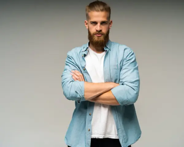 Uomo con barba e capelli biondi, camicia azzurra aperta su t-shirt bianca, braccia conserte. Look trasandato ma curato.