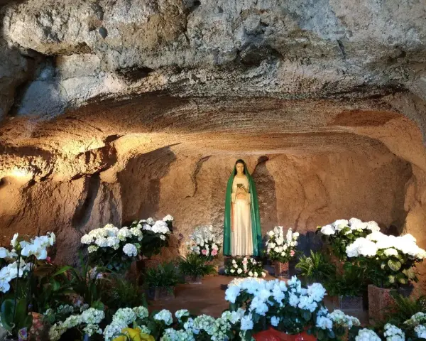 A statue of Mary stands in a grotto, surrounded by lush white flowers. The cave walls are illuminated, creating a serene atmosphere, reminiscent of the three fountains of grace.
