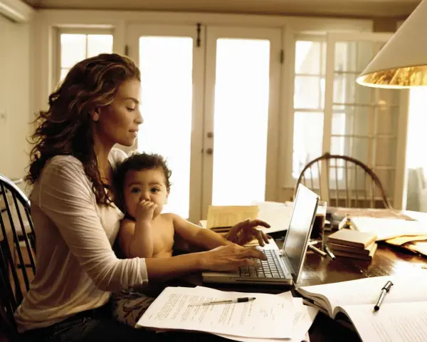 Una mamma lavoratrice con il suo bambino in grembo, mentre digita al computer tra libri e fogli.