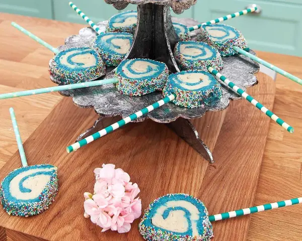 Blå och vita cake pops med strössel på ett silverfärgat fat. Vissa ligger på ett träunderlag med rosa blommor.