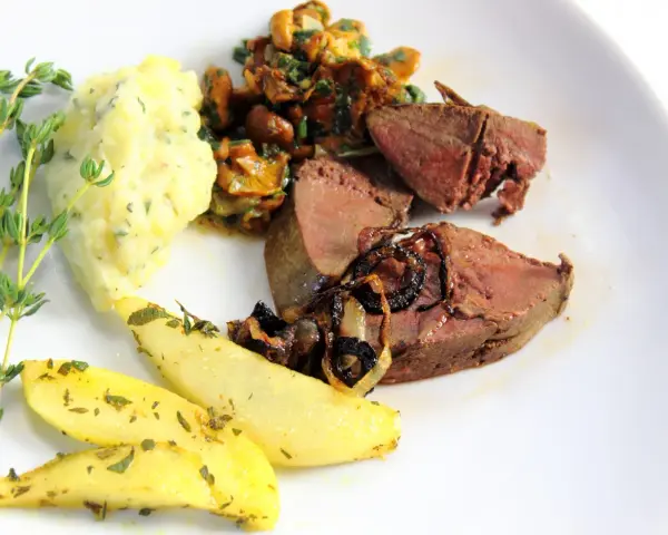 Rehleber zubereiten: zarte Scheiben Rehleber mit gebratenen Zwiebeln, Kartoffelpüree, gebratenen Kartoffelspalten und frischen Kräutern auf einem weißen Teller.