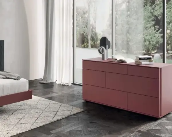 Cassettiera camera da letto moderna color bordeaux, con sei cassetti, accanto a un letto e una grande finestra.