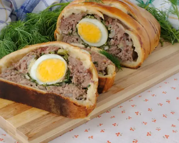 Une tranche de pâté de Pâques, garnie d'un œuf dur, est présentée sur une planche en bois, avec de l'aneth frais.