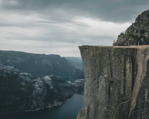Człowiek w żółtej kurtce stoi na krawędzi skalnego klifu w Norwegii, podziwiając majestatyczny fiord i góry.