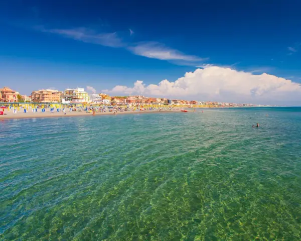 Słoneczna plaża w Rzymie, pełna kolorowych parasoli i ludzi kąpiących się w krystalicznie czystej wodzie.