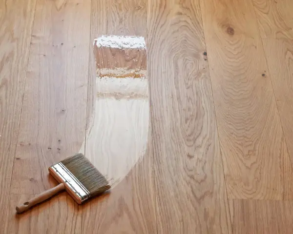 Breite Pinsel mit Holzgriff trägt Lack auf Parkett auf. Der Prozess, das Parkett zu versiegeln, zeigt den Unterschied zwischen unbehandeltem und frisch lackiertem Holz.