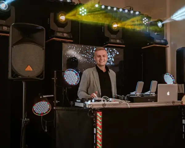 Uśmiechnięty dj na 18. urodziny, gotowy do rozkręcenia imprezy. W tle kolorowe światła i sprzęt nagłośnieniowy.