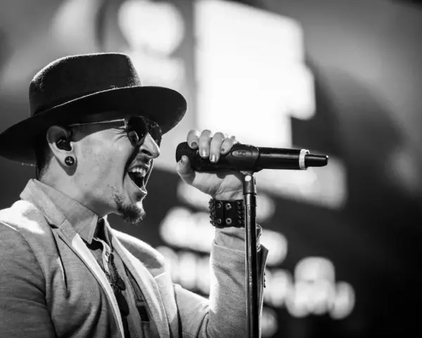 Wokalista Linkin Park, Chester Bennington, w kapeluszu i okularach przeciwsłonecznych, śpiewa do mikrofonu z pasją.