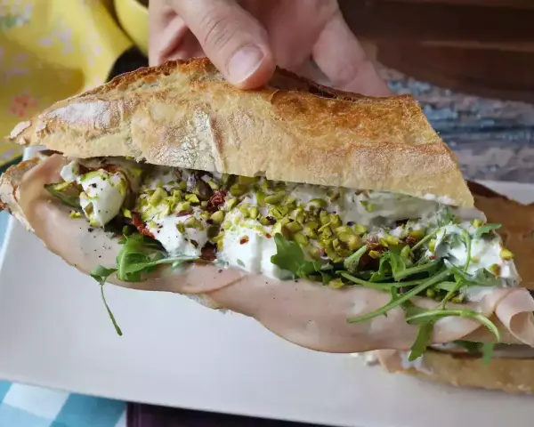 Bocadillo de pan rústico relleno de mortadela, burrata cremosa, rúcula, pistachos y tomates secos.