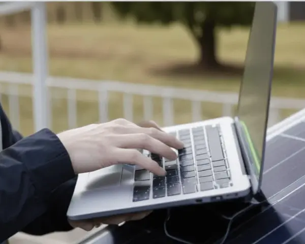 Praca nad projektem fotowoltaiki: ręce na klawiaturze laptopa, obok panel słoneczny.