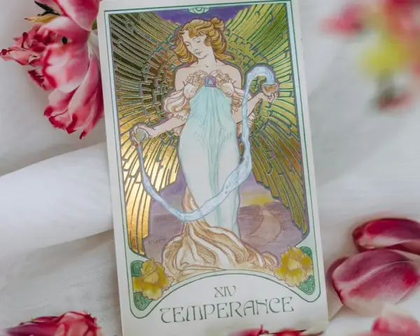 La carta de la templanza tarot, con su figura etérea vertiendo agua, rodeada de pétalos de tulipán rosa y blanco, evoca serenidad y equilibrio.