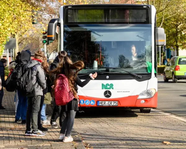 Ein Schulbus mit Warnblinker hält an einer Haltestelle. Schüler warten auf dem Gehweg, um einzusteigen.