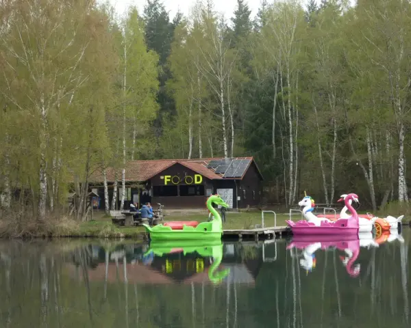 Bunte Tretboote in Form von Schwänen und Einhörnern liegen am Ufer des Bruchsee Duingen. Ein Gebäude mit der Aufschrift "FOOD" und Solaranlagen steht im Hintergrund.