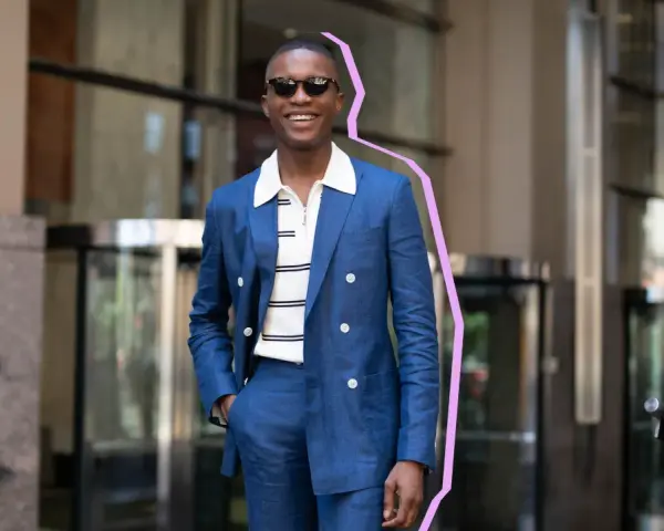 Hombre sonriente con traje azul y camiseta blanca a rayas, gafas de sol, posando con la mano en el bolsillo.