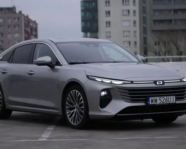 Srebrny Bestune B70 z nowoczesnymi światłami LED zaparkowany na betonowym placu, w tle budynki mieszkalne.