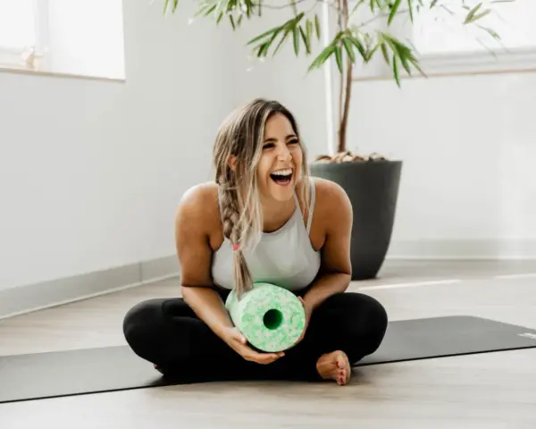 Lachende Frau mit Faszienrolle auf Yogamatte. Sie strahlt Lebensfreude aus und zeigt, wie man das positive sehen kann, auch nach dem Training.