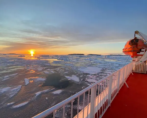 A **Greenland ferry** navigates icy waters at sunset. An orange lifeboat hangs ready on the ship's deck.