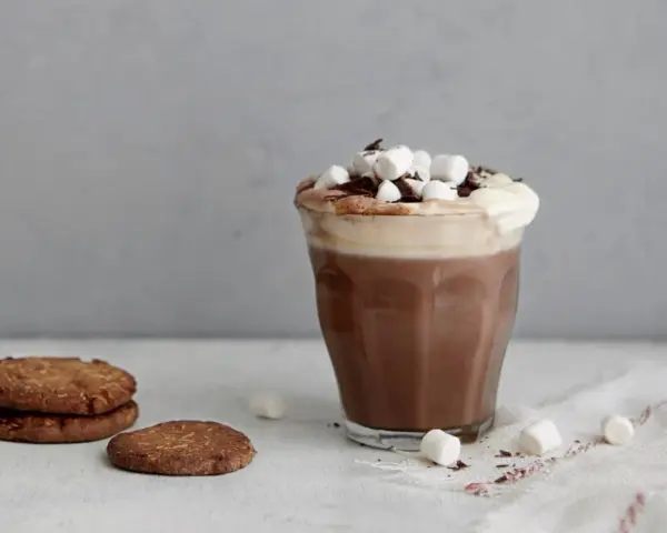 En härlig varm choklad med choklad, toppad med marshmallows och chokladspån. Bredvid står några chokladkakor.