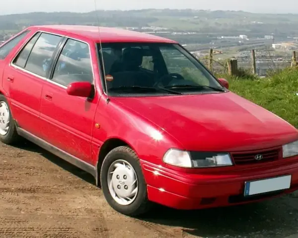 Czerwony Hyundai Pony z lat 90. stoi na polnej drodze, z widokiem na dolinę i miasto w tle.