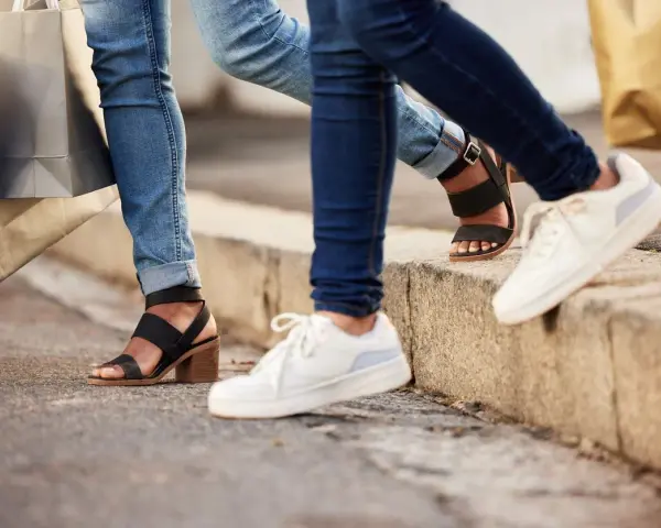 Dwie osoby w jeansach i modnych butach, z torbami na zakupy w rękach, idą chodnikiem. Jedna ma czarne sandały na obcasie, druga białe sneakersy.