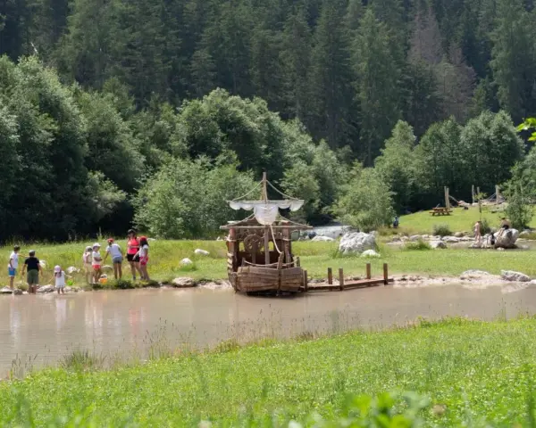 Bambini giocano vicino a un galeone di legno in un lago, mentre altri si rilassano sull'erba. Perfetto per le **san vigilio di marebbe passeggiate** estive.