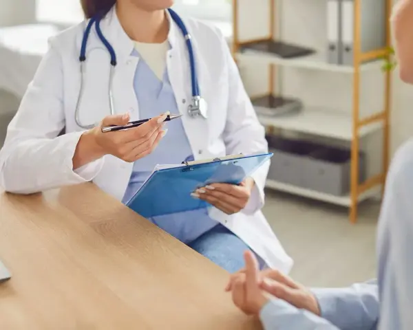 Lekarka w białym fartuchu z niebieskim stetoskopem rozmawia z pacjentem, trzymając teczkę.