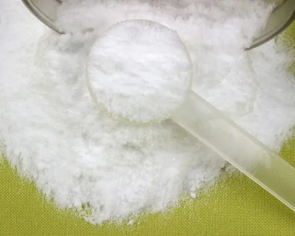 Biały proszek, prawdopodobnie l-glutamina, rozsypany na zielonym tle, z plastikową miarką.