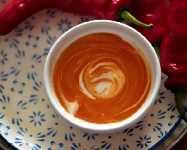 Crema di peperoni vellutata, con un vortice di panna, accanto a peperoni rossi freschi su un piatto decorato.