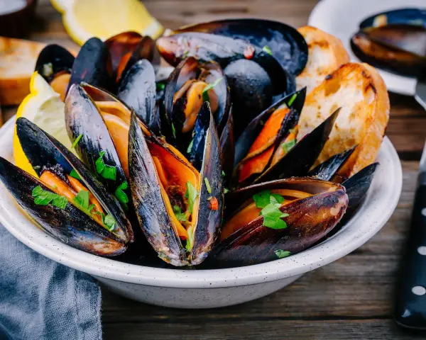 Mejillones en salsa, servidos en un cuenco blanco con perejil fresco, rodajas de limón y pan tostado.