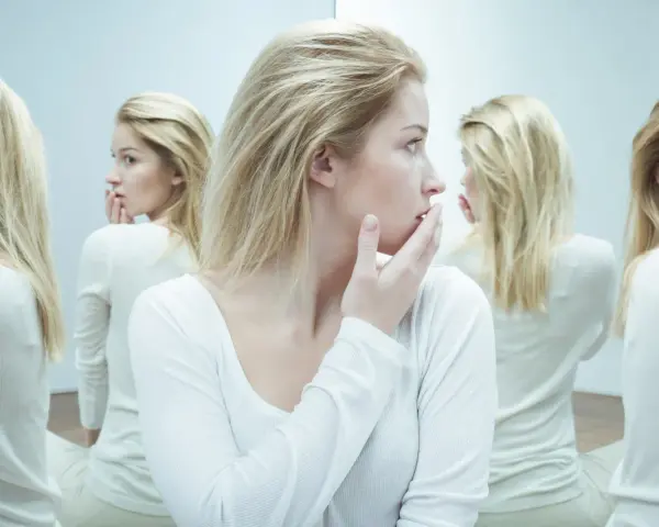 Kobieta z blond włosami w białej bluzce, otoczona swoimi odbiciami w lustrze.