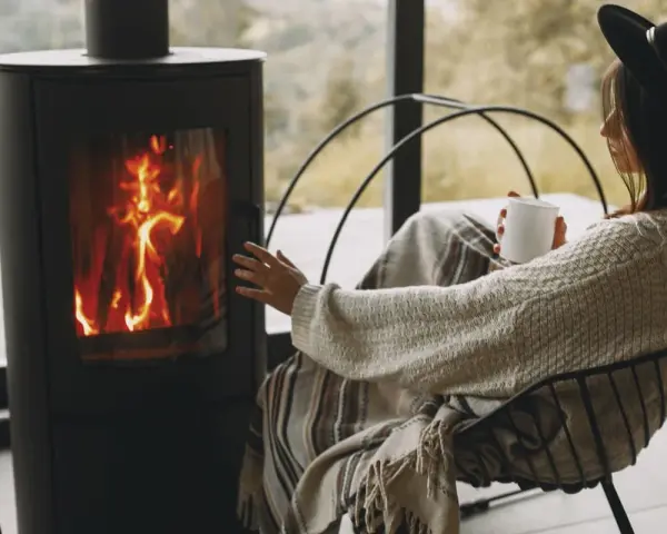 Donna avvolta in una coperta, con una tazza in mano, si scalda vicino a una stufa a legna, un modo accogliente per riscaldare la casa senza termosifoni.
