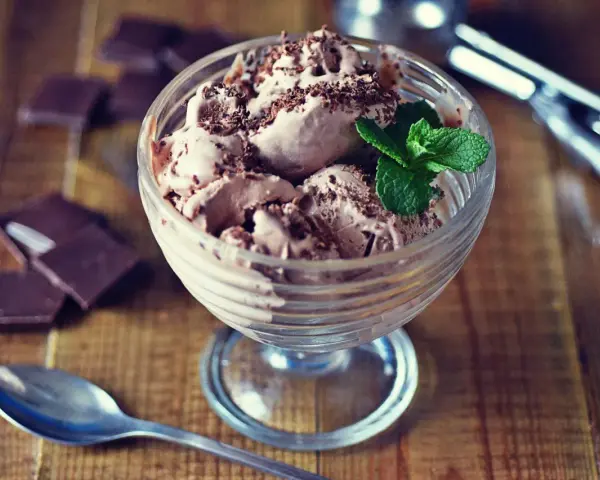 Pyszne lody nuii w pucharku, posypane czekoladą i udekorowane miętą. Obok leżą kawałki czekolady i łyżeczka.