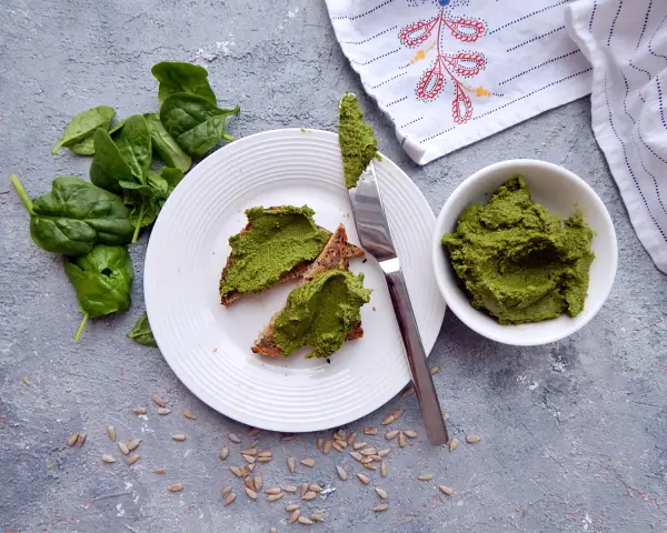 Pesto ze szpinaku do słoików na chlebie z pestkami słonecznika. Świeży szpinak obok talerza.