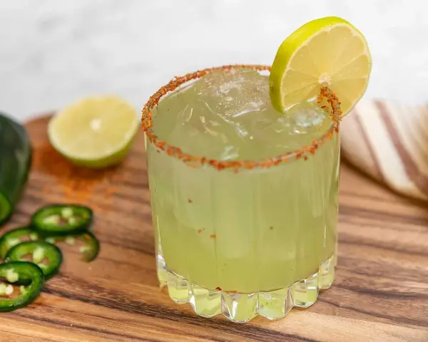 En glas med en kryddig margarita, garnerad med lime och chilipulver. Bredvid ligger jalapeños och limeklyftor.