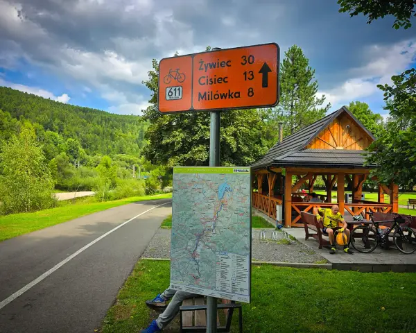 Znak drogowy dla rowerzystów na szlaku Velo Sola. Wskazuje kierunki i odległości do Żywca, Ciśca i Milówki. Obok mapa szlaku.