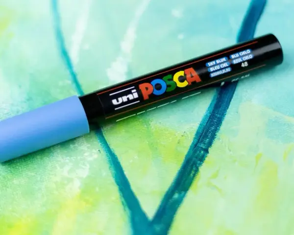 En ljusblå Posca-tuschpenna ligger på en abstrakt målning i gröna och blå nyanser. Perfekt för att måla med tuschpennor.
