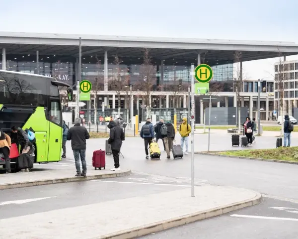 Ludzie z bagażami wsiadają do zielonego autokaru przed nowoczesnym budynkiem, przypominającym lotniska w Berlinie.