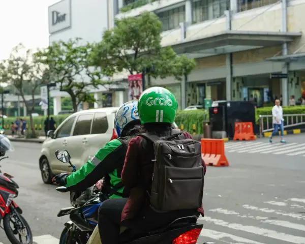 Zwei Personen auf einem Roller, der Fahrer trägt einen grünen Helm mit dem "Grab" Logo. Sie sind unterwegs in Thailand, um mit der Grab App Kunden zu transportieren.