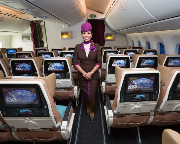 Uśmiechnięta stewardessa Etihad Airways w brązowym mundurku stoi w rzędzie foteli. Ekraniki pokazują astronautę. Dobre etihad airways opinie.