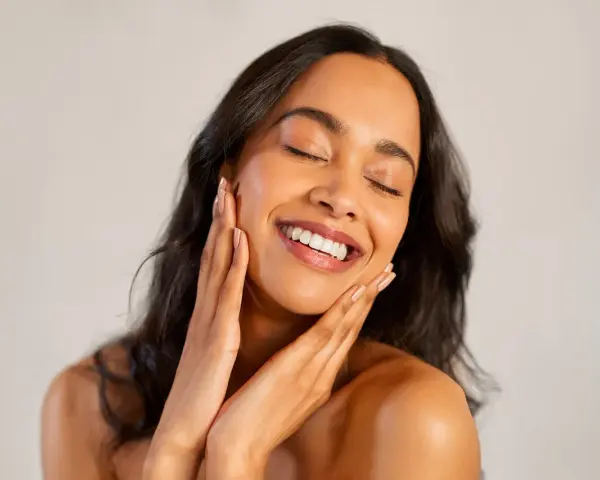 Une femme au sourire radieux caresse son visage, révélant une peau glowy éclatante de santé.