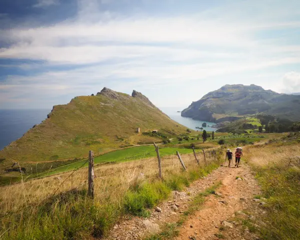 Two hikers with backpacks walk on a dirt path through a grassy landscape with mountains and the sea in the background.