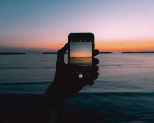 Dłoń trzyma smartfon, na ekranie którego widać piękny zachód słońca nad morzem. Idealny kadr, by nauczyć się, jak fotografować zachód słońca.