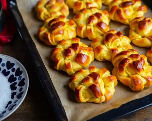Nybakade lussebullar med kesella, gyllene och redo att avnjutas. En skål med socker och en röd duk anas i bakgrunden.