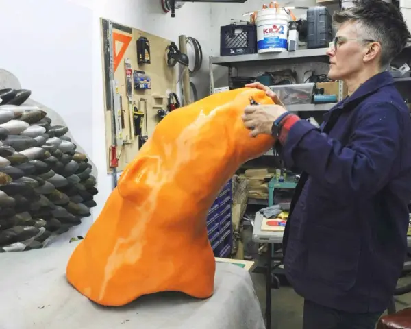 Artist applies finishing touches to a large, orange, 3D printed object, part of a project creating large objects.