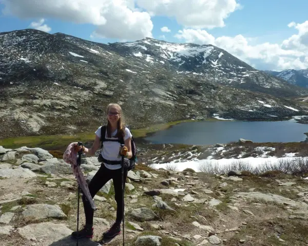Jeune randonneuse souriante avec bâtons, face à une **mare** et des montagnes enneigées. La **difficulté** du sentier **nord** est palpable.