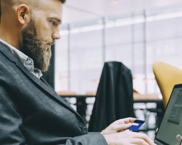 Hombre con barba y traje usa tarjeta de crédito en portátil. Busca cuánto se paga la hora extra en España.